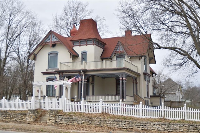 victorian-style house with a fenced front yard, covered porch, a chimney, and a balcony