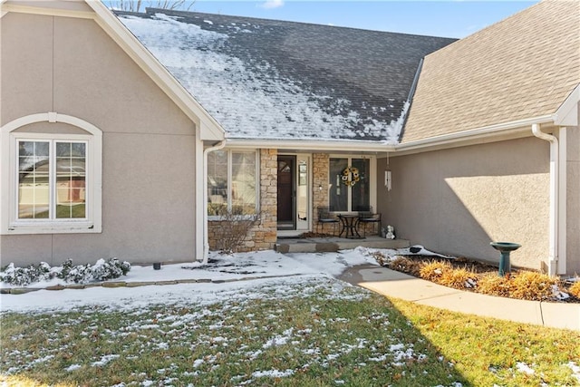 snow covered property entrance with stucco siding, a shingled roof, a porch, an attached garage, and stone siding