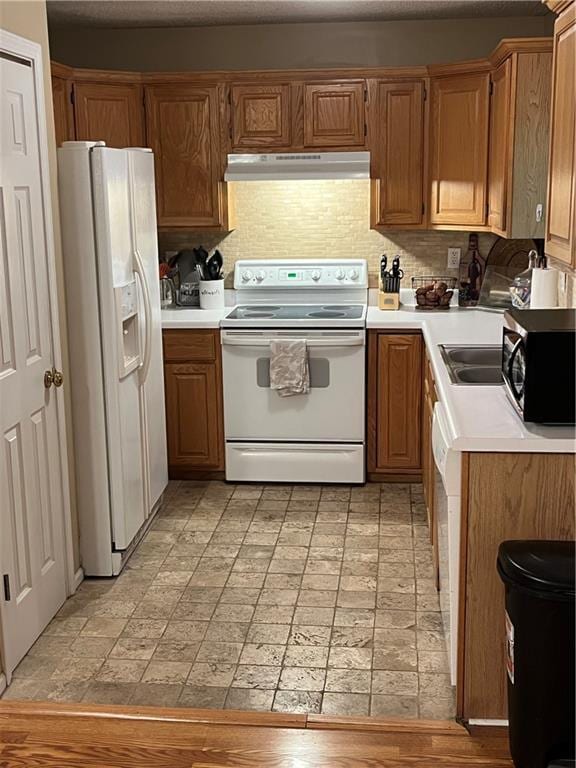 kitchen with under cabinet range hood, white appliances, light countertops, backsplash, and brown cabinets