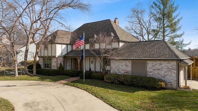 tudor-style house with brick siding, a chimney, a shingled roof, a front yard, and driveway