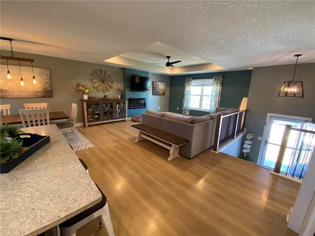 living area featuring baseboards, wood finished floors, a tray ceiling, a textured ceiling, and ceiling fan with notable chandelier