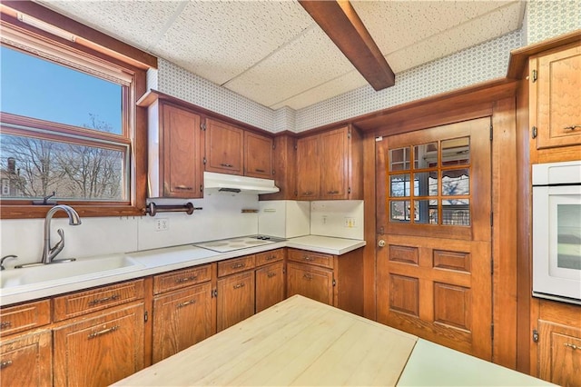 kitchen with under cabinet range hood, white appliances, a sink, light countertops, and brown cabinets