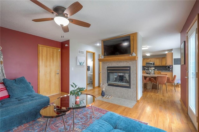 living room with visible vents, a brick fireplace, light wood-style flooring, and a ceiling fan