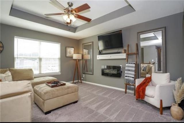 living area featuring carpet floors, a raised ceiling, a glass covered fireplace, ceiling fan, and baseboards