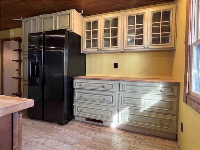 kitchen with light wood finished floors, black fridge with ice dispenser, glass insert cabinets, cream cabinets, and light countertops