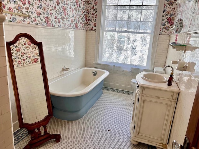 bathroom with tile walls, a soaking tub, wainscoting, vanity, and wallpapered walls