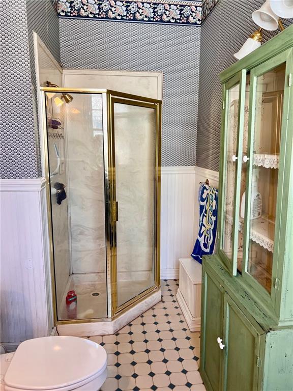 bathroom featuring a wainscoted wall, wallpapered walls, toilet, a shower stall, and tile patterned floors