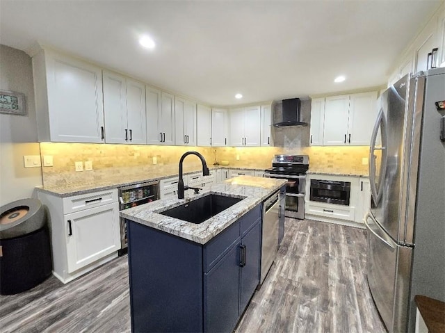 kitchen featuring wall chimney exhaust hood, a kitchen island with sink, stainless steel appliances, white cabinetry, and a sink