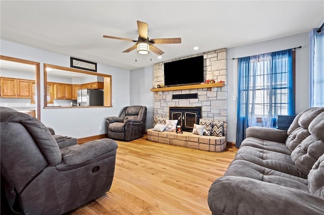 living room featuring light wood finished floors, a stone fireplace, baseboards, and a ceiling fan