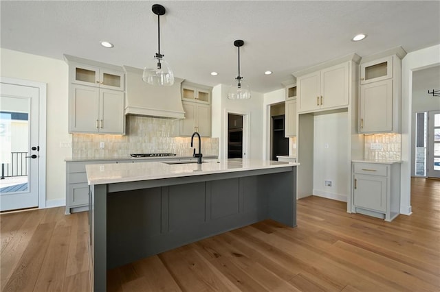 kitchen featuring pendant lighting, a center island with sink, light countertops, glass insert cabinets, and a sink