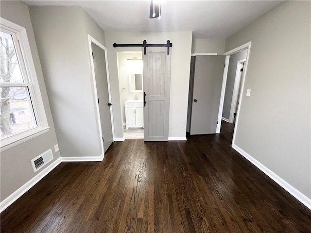 unfurnished bedroom with a barn door, dark wood finished floors, visible vents, baseboards, and ensuite bath