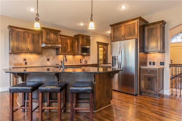 kitchen featuring a sink, stainless steel appliances, dark wood-type flooring, under cabinet range hood, and a kitchen bar