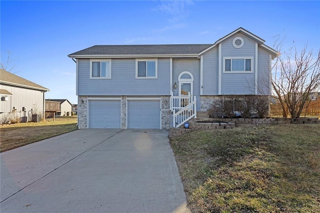 raised ranch with stone siding, central AC, concrete driveway, a front yard, and a garage