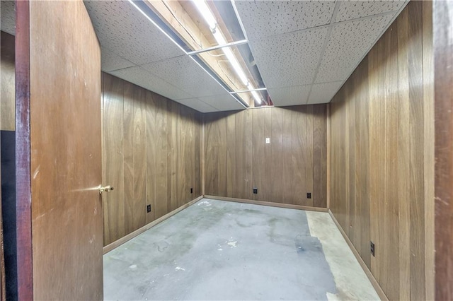 empty room with concrete floors, wood walls, a drop ceiling, and baseboards