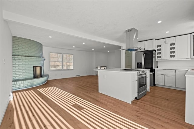 kitchen with open floor plan, appliances with stainless steel finishes, island range hood, light wood finished floors, and a brick fireplace