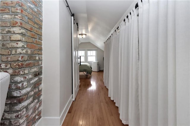 hall with a barn door, wood finished floors, brick wall, and vaulted ceiling