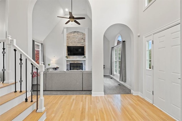 foyer entrance featuring stairway, ceiling fan, a stone fireplace, wood finished floors, and high vaulted ceiling