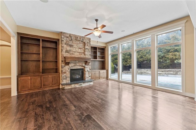 unfurnished living room with built in features, baseboards, dark wood finished floors, a ceiling fan, and a stone fireplace