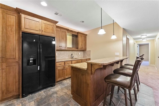 kitchen featuring tasteful backsplash, black refrigerator with ice dispenser, brown cabinetry, a sink, and a kitchen bar