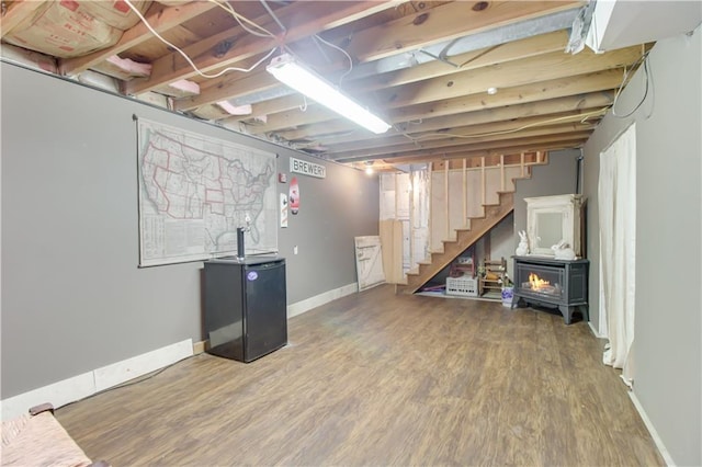 unfinished basement featuring freestanding refrigerator, a wood stove, wood finished floors, baseboards, and stairs