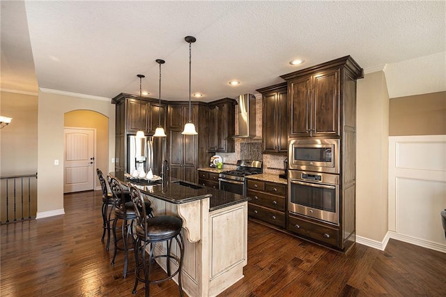 kitchen with an island with sink, wall chimney exhaust hood, appliances with stainless steel finishes, dark stone countertops, and decorative light fixtures