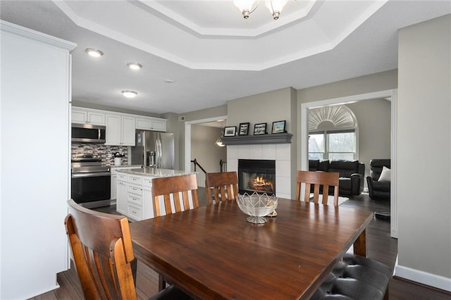 dining space with a notable chandelier, dark wood-style flooring, a fireplace, baseboards, and a raised ceiling