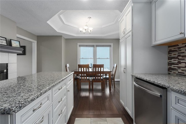 kitchen featuring a raised ceiling, backsplash, stainless steel dishwasher, an inviting chandelier, and light stone countertops