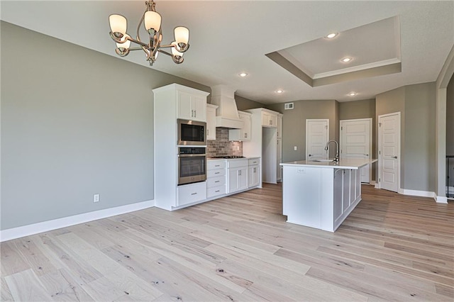 kitchen with a raised ceiling, a kitchen island with sink, custom exhaust hood, built in microwave, and stainless steel oven