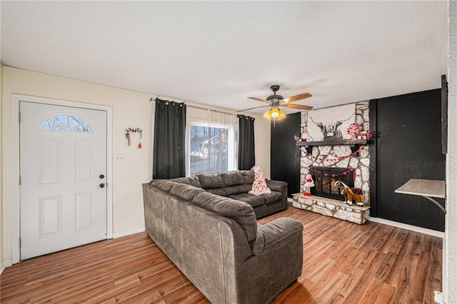 living area featuring ceiling fan, a textured ceiling, a stone fireplace, wood finished floors, and baseboards