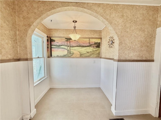 unfurnished dining area featuring a wainscoted wall, light tile patterned floors, arched walkways, and wallpapered walls