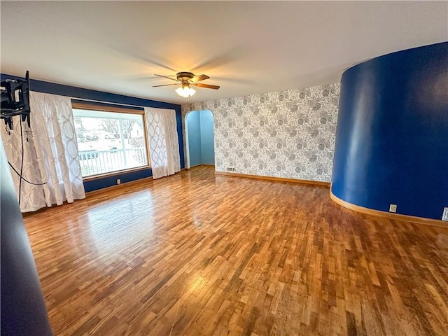 spare room featuring arched walkways, ceiling fan, wood finished floors, baseboards, and wallpapered walls