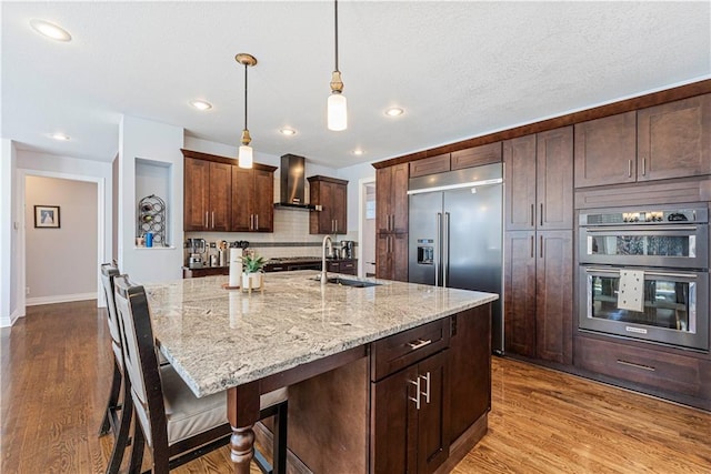 kitchen featuring stainless steel appliances, dark wood-type flooring, a sink, wall chimney range hood, and light stone countertops