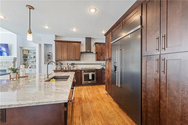 kitchen featuring light wood finished floors, decorative light fixtures, stainless steel appliances, wall chimney range hood, and a sink