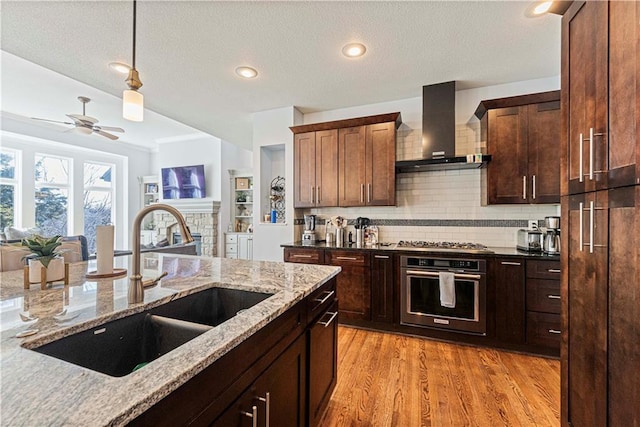 kitchen featuring light wood finished floors, stainless steel gas stovetop, wall oven, a sink, and wall chimney exhaust hood