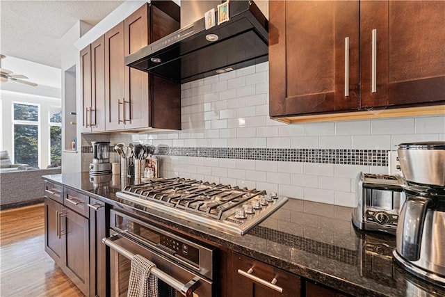 kitchen with decorative backsplash, appliances with stainless steel finishes, a ceiling fan, wall chimney range hood, and light wood-type flooring