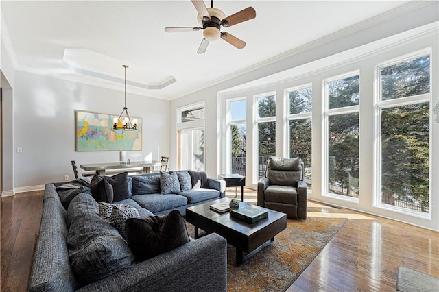 living area with a raised ceiling, ornamental molding, wood finished floors, baseboards, and ceiling fan with notable chandelier