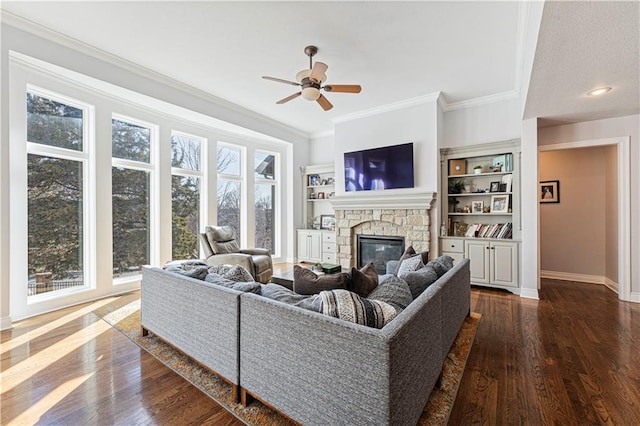 living area with dark wood-style floors, crown molding, a ceiling fan, a stone fireplace, and baseboards