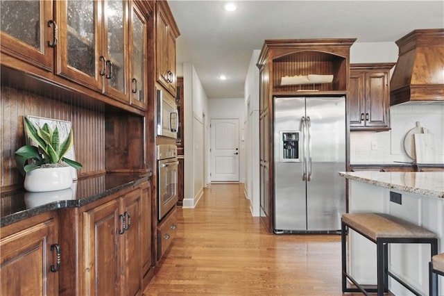 kitchen featuring appliances with stainless steel finishes, light wood-style floors, stone counters, custom range hood, and glass insert cabinets