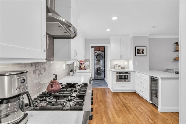 kitchen featuring wall chimney exhaust hood, wine cooler, stacked washer / drying machine, oven, and white cabinetry