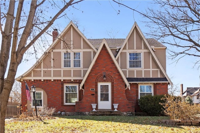 tudor house featuring stucco siding, fence, roof with shingles, brick siding, and a chimney