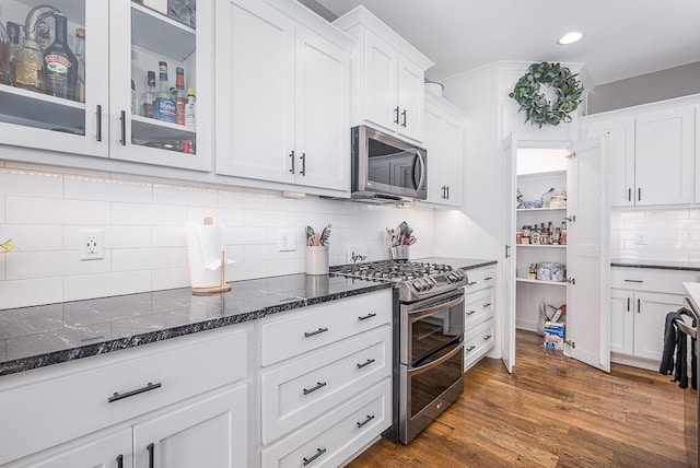 kitchen featuring dark wood-style floors, stainless steel appliances, dark stone counters, white cabinets, and glass insert cabinets