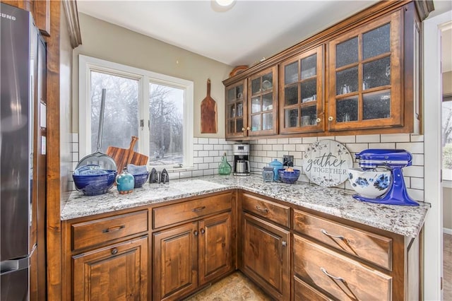 kitchen with tasteful backsplash, glass insert cabinets, freestanding refrigerator, and light stone countertops