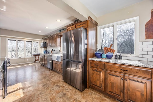 kitchen featuring backsplash, light stone counters, brown cabinetry, stainless steel appliances, and a sink