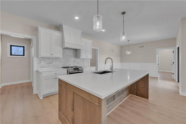 kitchen with stainless steel gas stove, custom range hood, a kitchen island with sink, light countertops, and a sink