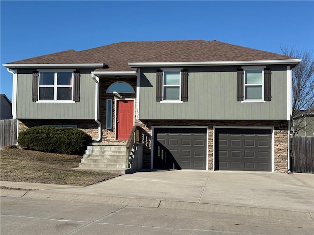 bi-level home featuring a garage, a shingled roof, fence, driveway, and stone siding