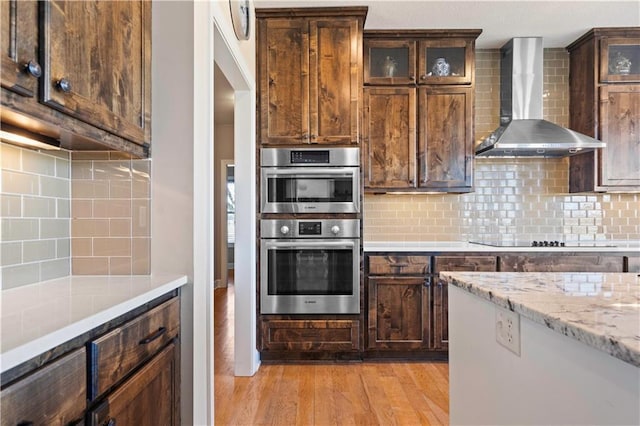 kitchen featuring black electric stovetop, wall chimney range hood, stainless steel double oven, and backsplash