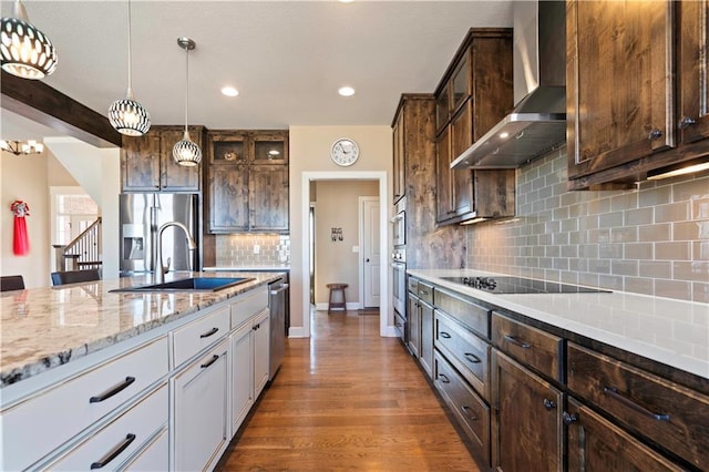 kitchen with wood finished floors, hanging light fixtures, stainless steel appliances, wall chimney range hood, and a sink