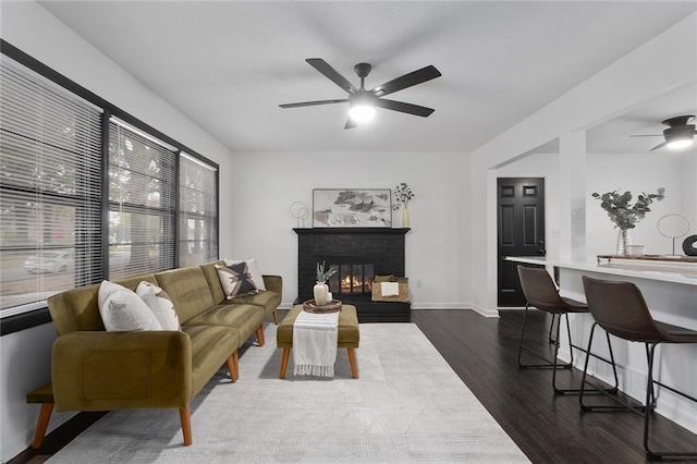 living area featuring a brick fireplace, baseboards, a ceiling fan, and dark wood-style flooring