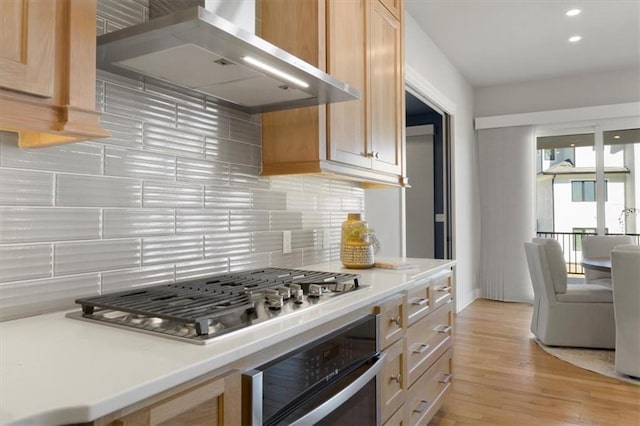 kitchen featuring light wood finished floors, light countertops, decorative backsplash, appliances with stainless steel finishes, and wall chimney exhaust hood