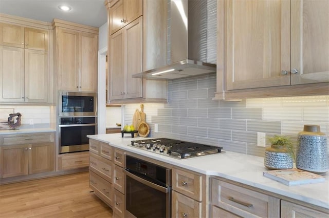 kitchen featuring light wood finished floors, stainless steel appliances, light countertops, light brown cabinetry, and wall chimney range hood